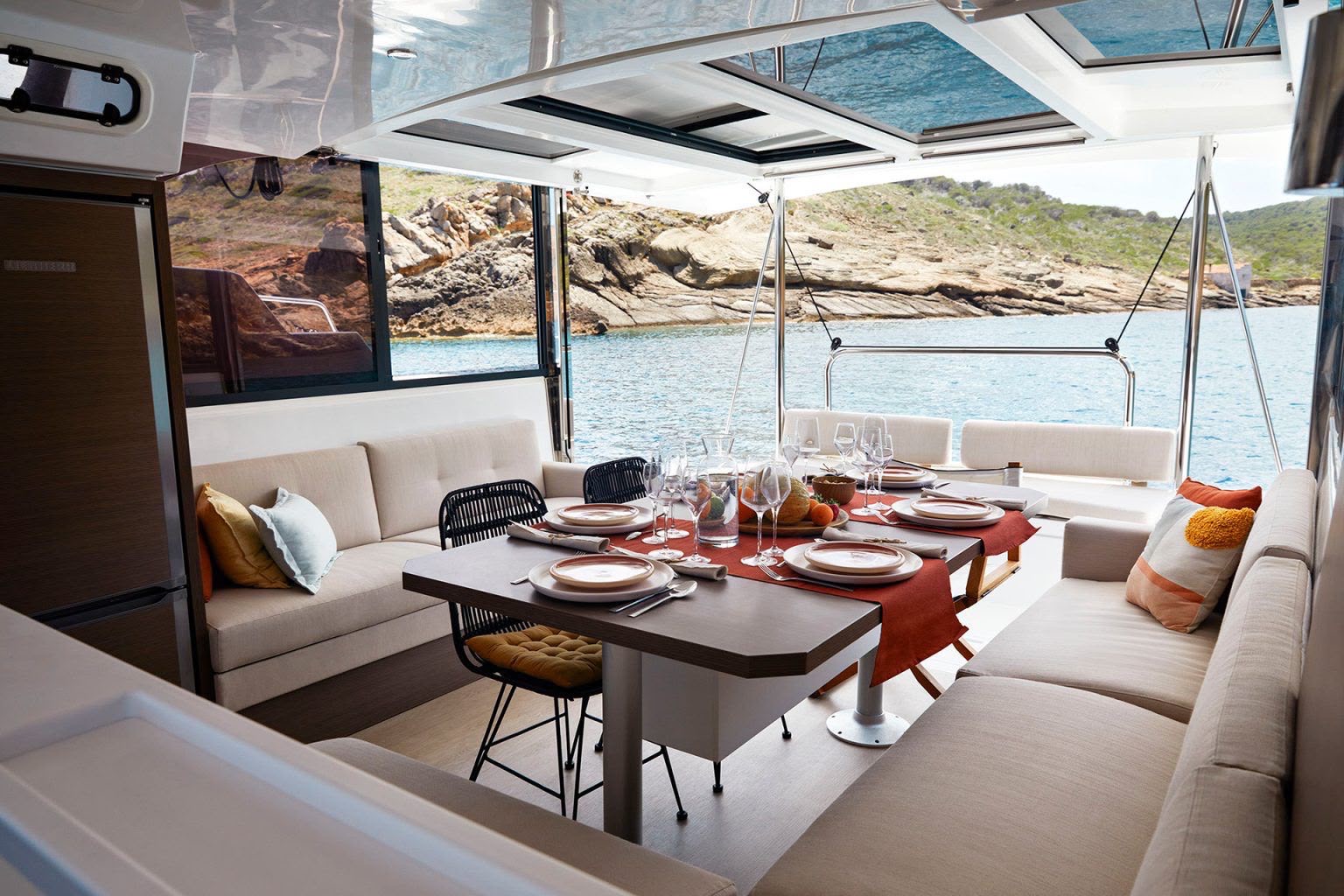 Zona exterior del catamarán con la mesa preparada y sofás, frente a una costa rocosa.